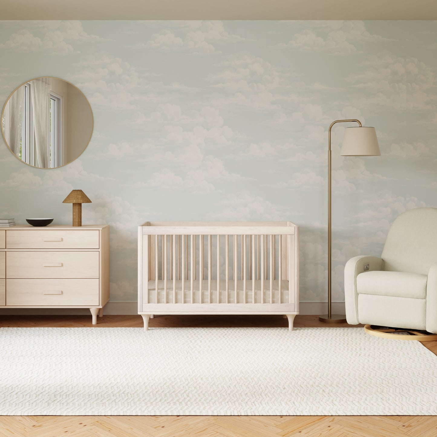 A minimal nursery features the Babyletto Lolly Convertible Crib, matching dresser, round mirror, modern floor lamp, table lamp, upholstered armchair, and soft rug on wooden floors. The wall is decorated with a pale cloud pattern.