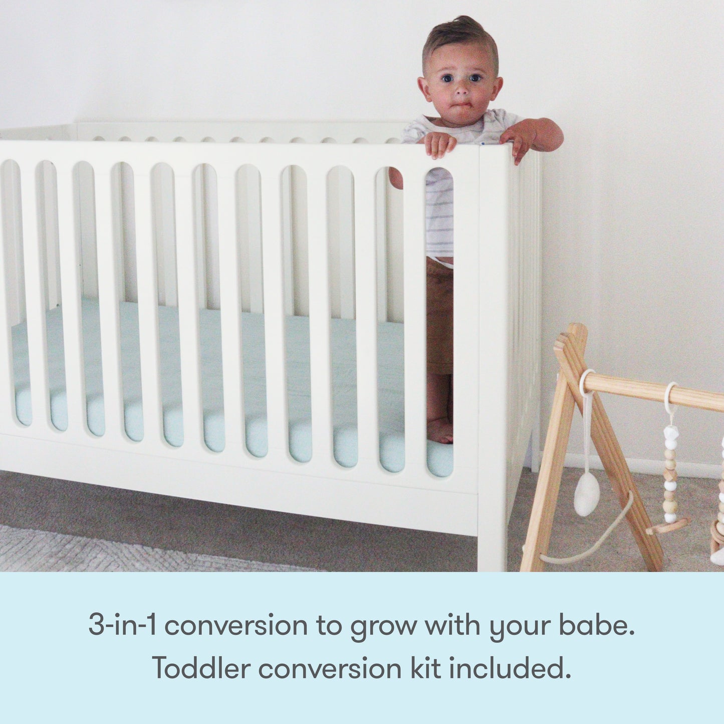 A toddler stands holding the rail of the white Newton Galileo Convertible Crib by Newton Baby. Next to it is a wooden baby gym with hanging toys. Text: 3-in-1 convertible crib grows with your child. Toddler bed conversion kit included.