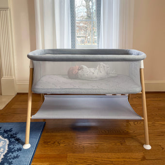 Sleeping baby in gray essential bassinet with full wood legs and storage shelf in a neutral room with a blue rug.