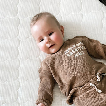 A baby in a brown sweatsuit lies comfortably on a Newton mattress, looking peaceful and relaxed.