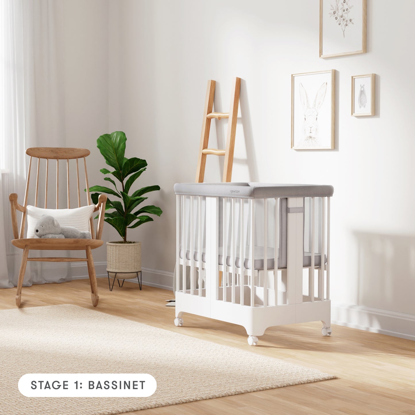 White color Newton Nest Convertible Crib in bassinet setting with mesh liner, in nursery next to rocking chair, with text "Stage 1: Bassinet"
