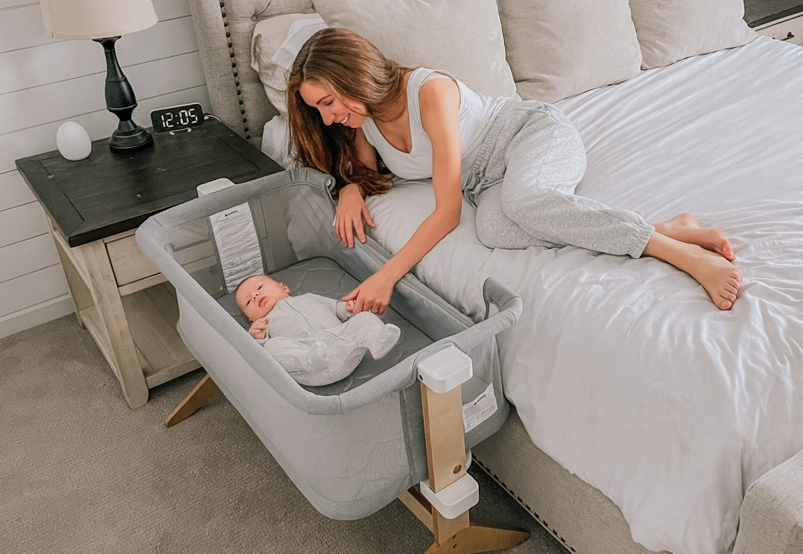 A joyful mother in a white and gray bedroom smiles as she interacts with her baby lying contently in a modern gray bassinet.