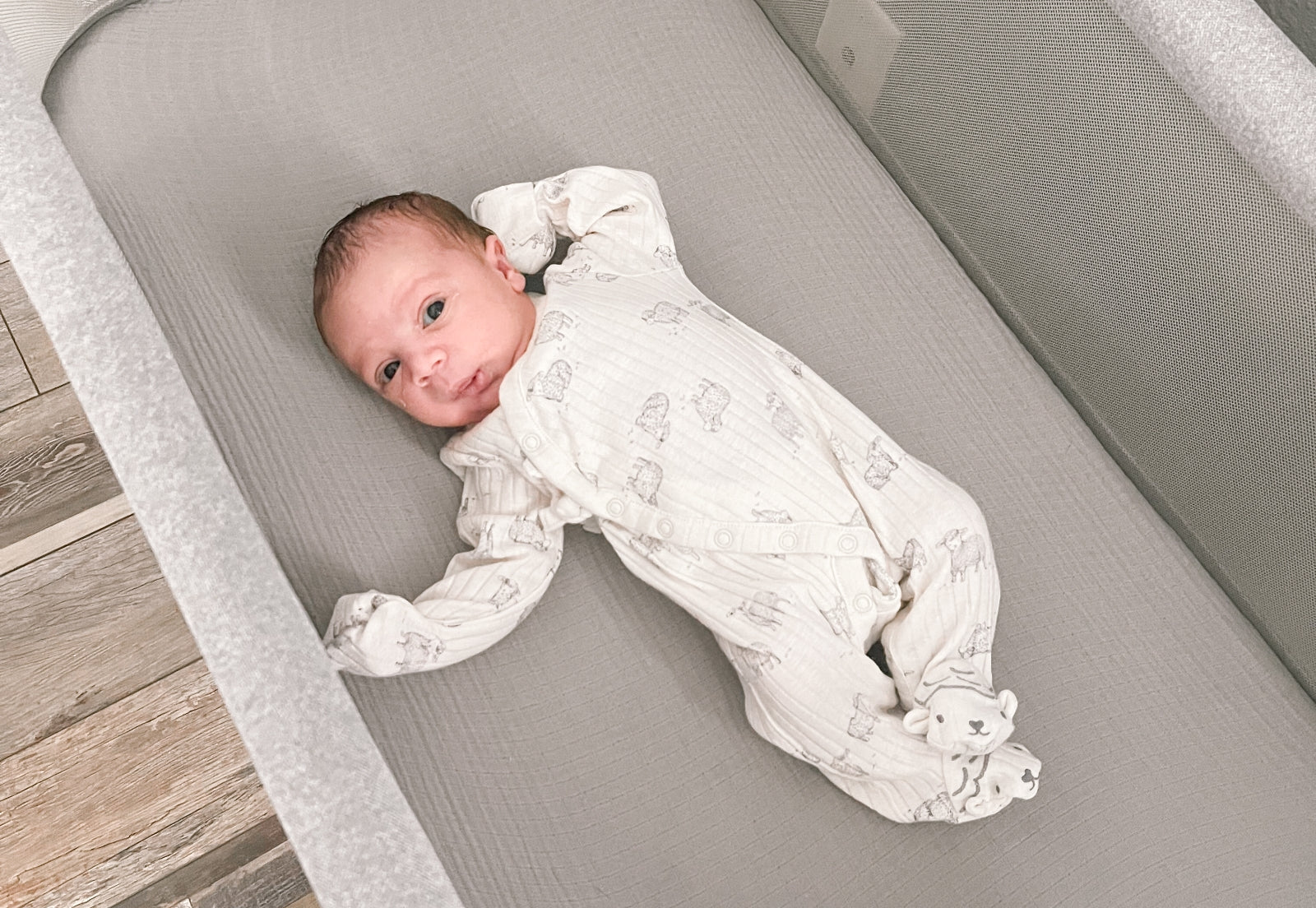ALT text: "A newborn baby dressed in a white onesie with animal prints lies comfortably in a gray, breathable Newton Baby crib mattress."