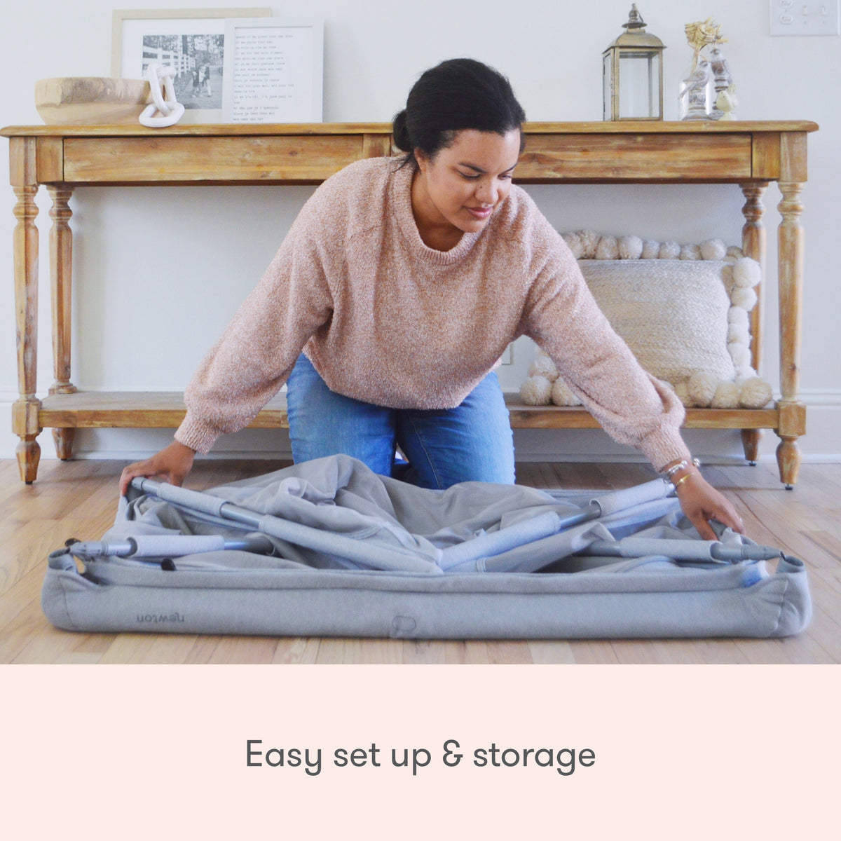 A woman kneels on a wooden floor, folding a gray Newton Baby Deluxe Travel Crib & Play Yard. A wooden console table with decor is behind her. Text reads: Easy set up & storage—perfect for any play yard or nursery.