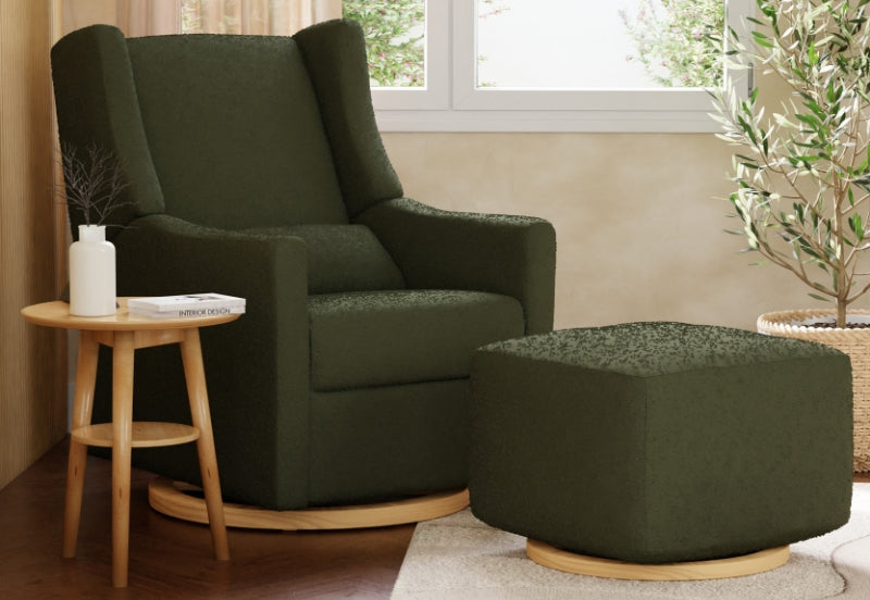 Cozy green nursery rocking chair and matching ottoman in a well-lit room with a small wooden table featuring a book titled "Interior Design," a white candle, and a decorative plant.