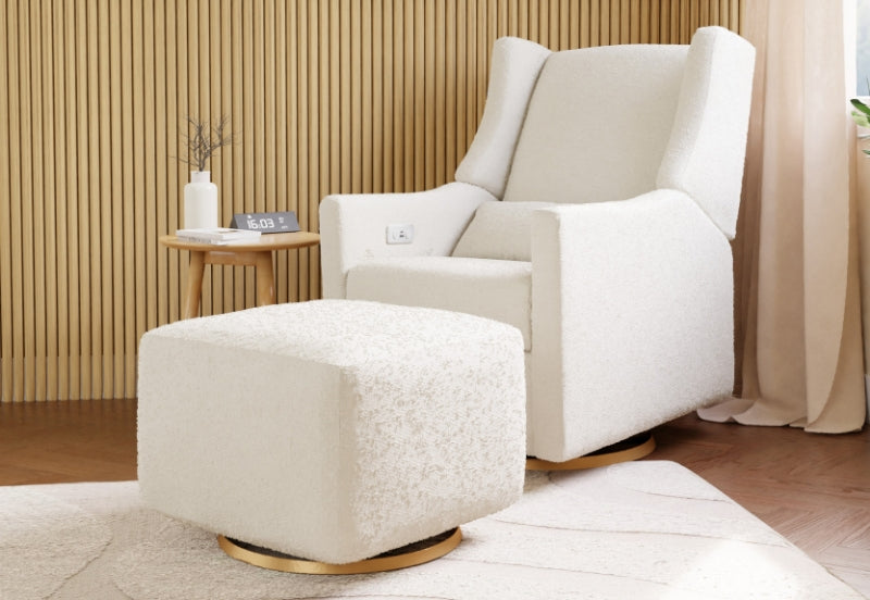 A cozy, modern nursery featuring a plush, white upholstered rocking chair and matching ottoman, set against a warm wooden panel backdrop with a small round table displaying a clock reading "16:03" and a candle.