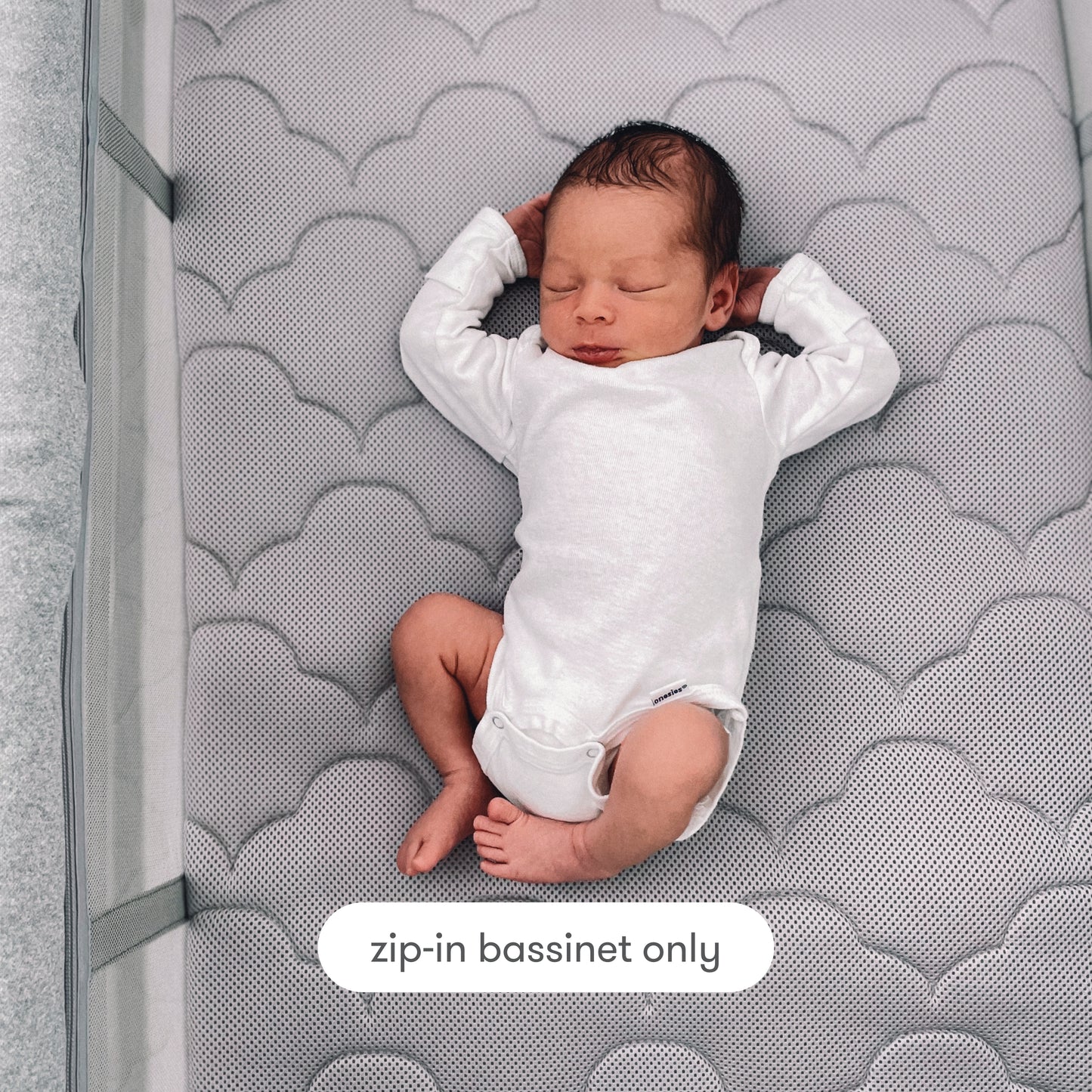 A newborn in a white onesie sleeps on a gray, cloud-patterned breathable mattress inside the Newton Baby Deluxe Travel Crib & Play Yard bassinet. At the bottom, a label reads “zip-in bassinet only.”.