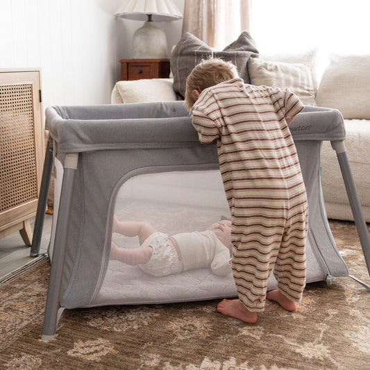 A toddler in striped pajamas stands by the Newton Baby Compact Travel Crib & Play Yard, watching a baby inside. The cozy living room features a couch, lamp, and soft lighting. 999-5101-GRY
