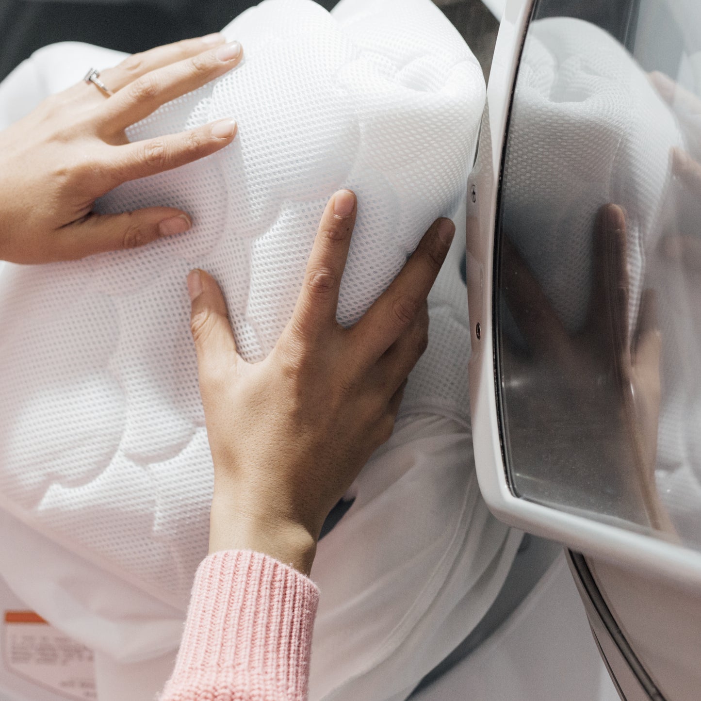 A person in a pink sweater places a Newton Baby Waterproof Crib Mattress Pad into a washing machine.