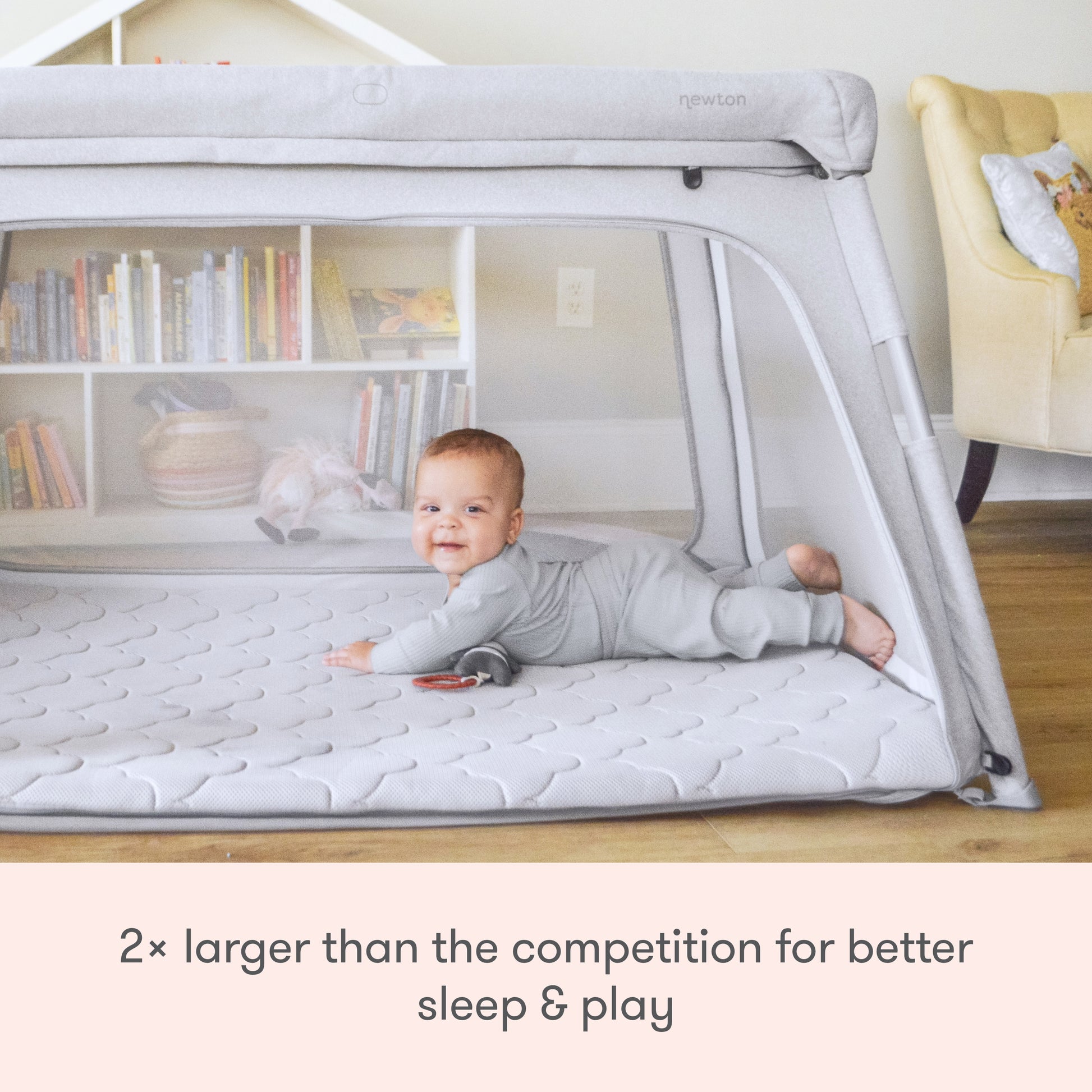A happy baby lies on their tummy inside the Newton Baby Deluxe Travel Crib & Play Yard, featuring breathable mesh sides and mattress. Toys sit nearby. Text reads: 2× larger than competitors for better sleep & play.