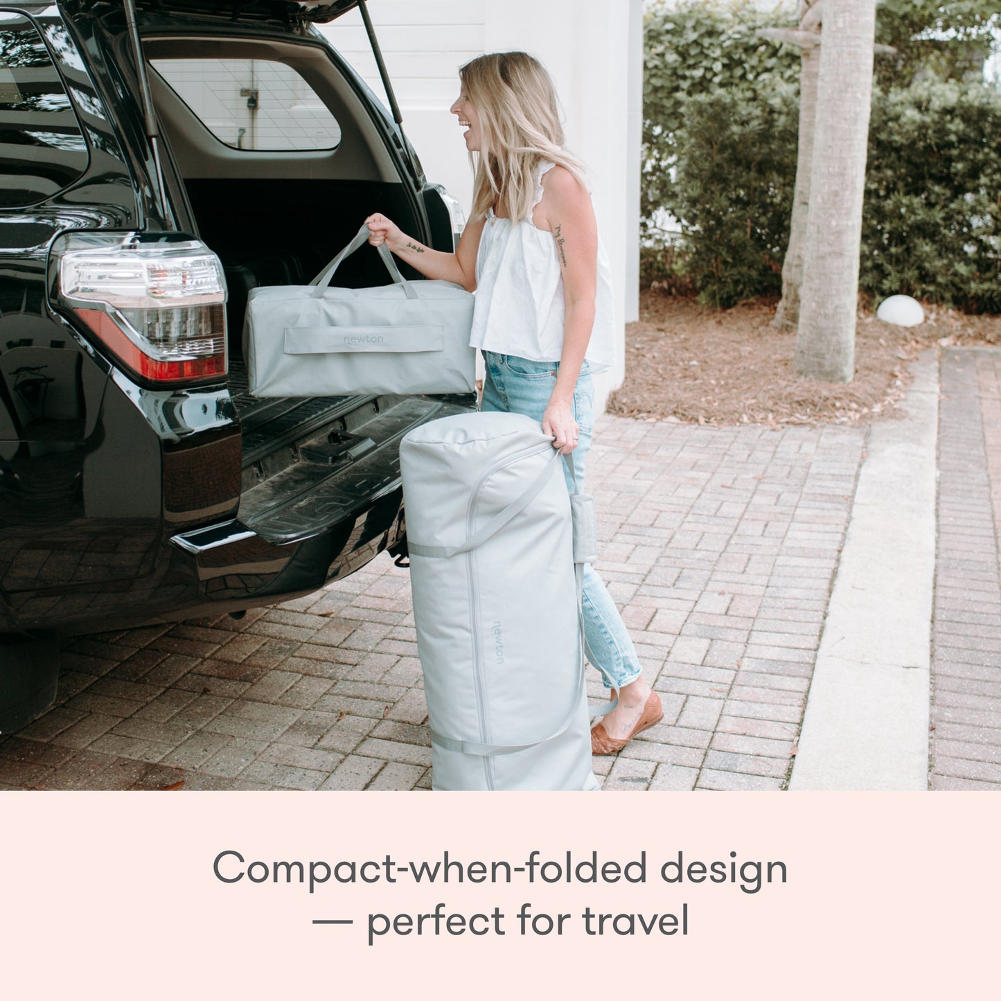 A woman loads the Newton Baby Deluxe Travel Crib & Play Yard, in light gray, into an SUV trunk while holding the matching duffel. Text: Compact-when-folded design with breathable mattress—perfect for travel.