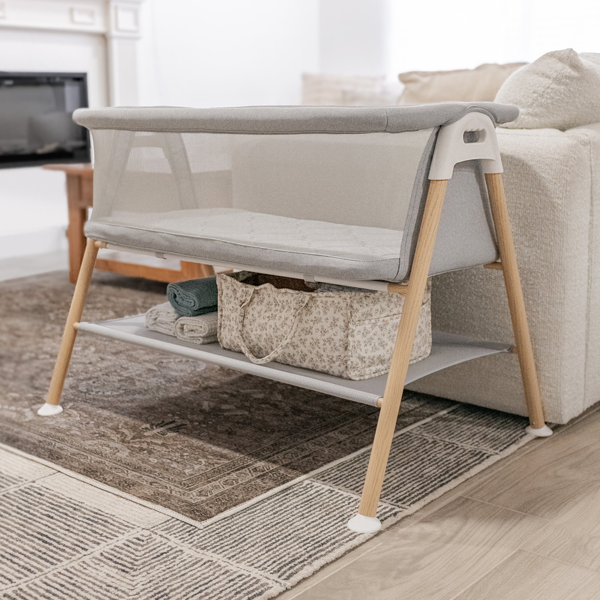 essential bassinet next to a couch in a living room, storage space below bassinet filled with sheets and other baby care essentials