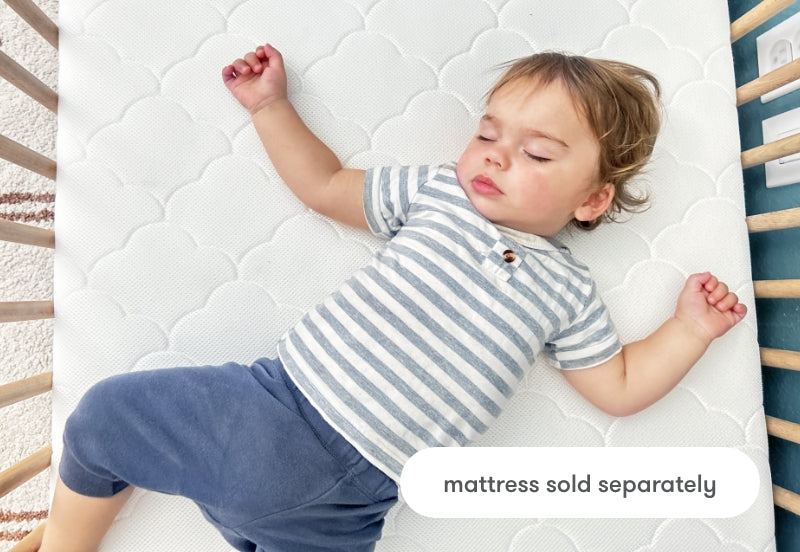 Toddler sleeping peacefully on a patterned white mattress, dressed in a striped shirt and blue shorts, with text stating "mattress sold separately".