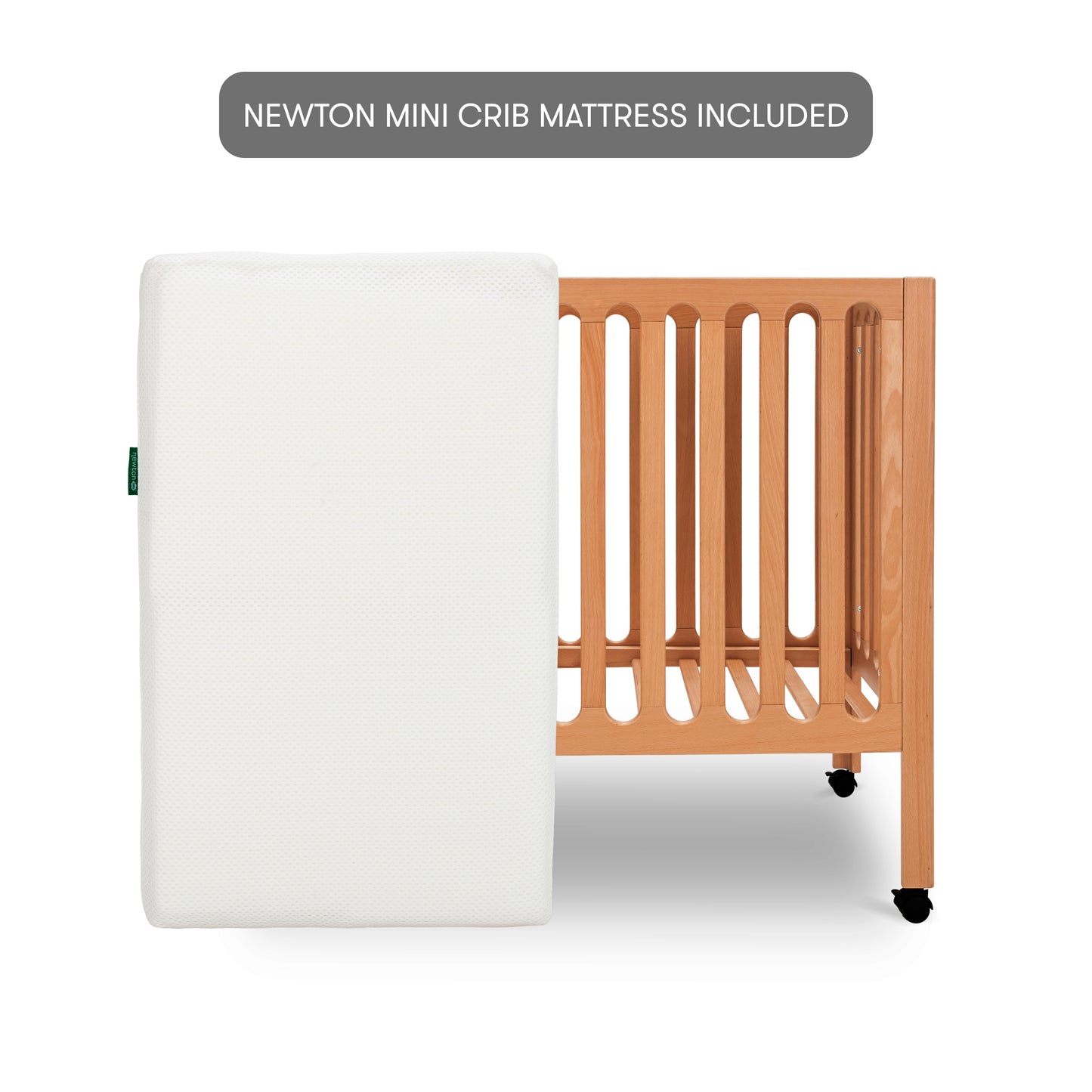 Modern wooden crib on wheels with mattress leaned up against crib, isolated on a white background. With text, "newton mini crib mattress included".
