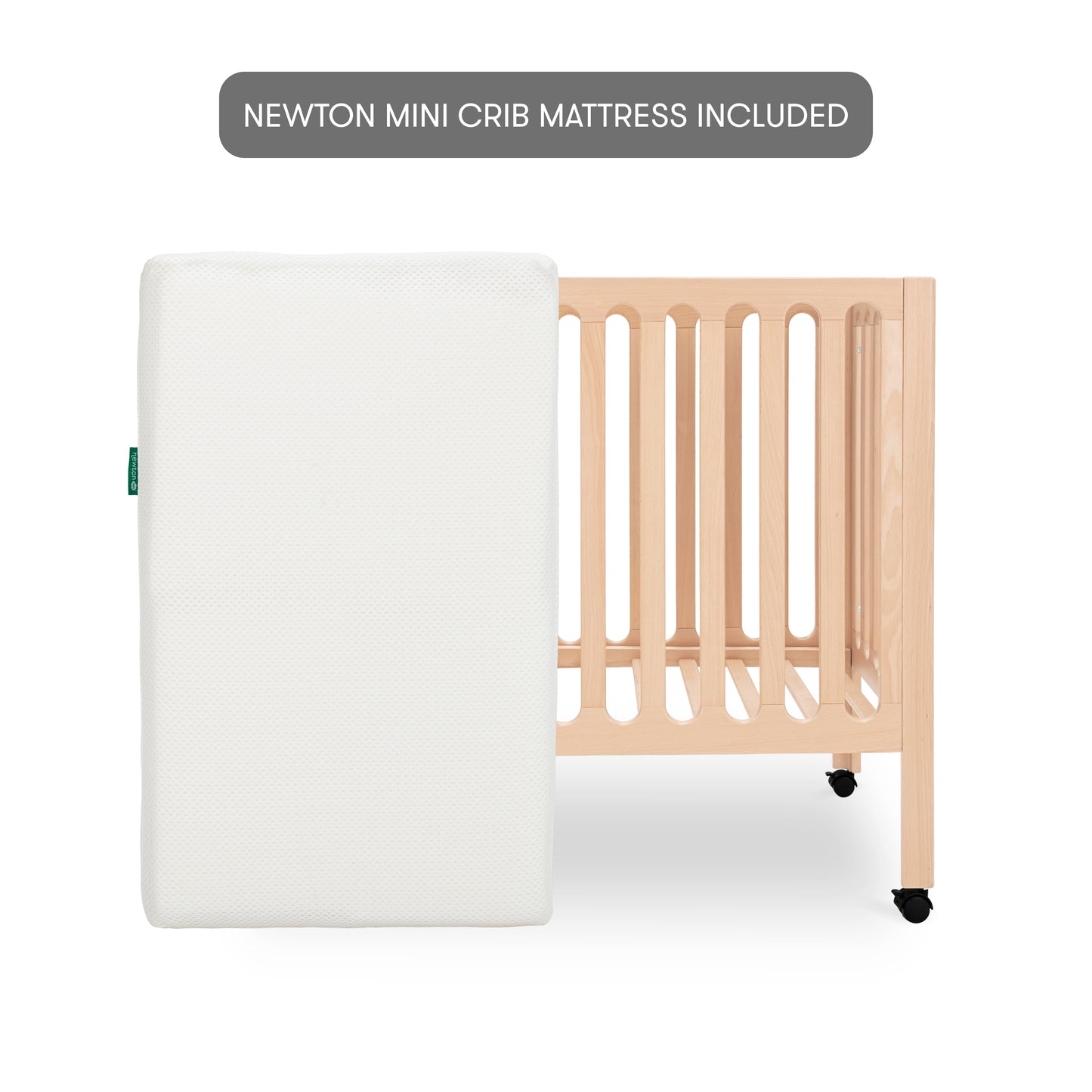 Modern wooden baby crib on wheels, featuring a white mattress leaned up against the crib with a minimalist design. With text "newton mini crib mattress included"