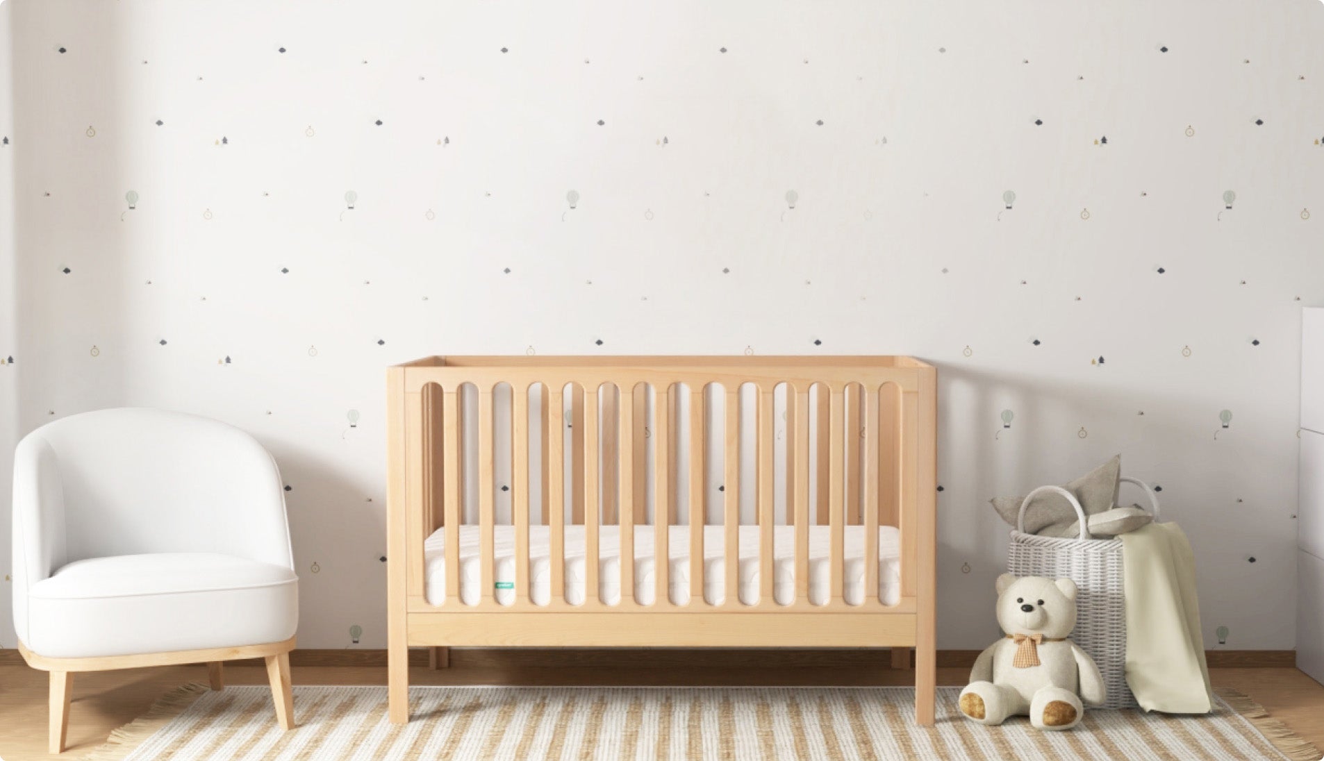 A modern nursery featuring a Newton convertible crib made of light wood with vertical slats, placed against a white wall with subtle balloon and cloud wallpaper. To the left is a white cushioned chair, and to the right is a white wicker basket with pillows, a cream-colored blanket, and a plush teddy bear on a striped rug.