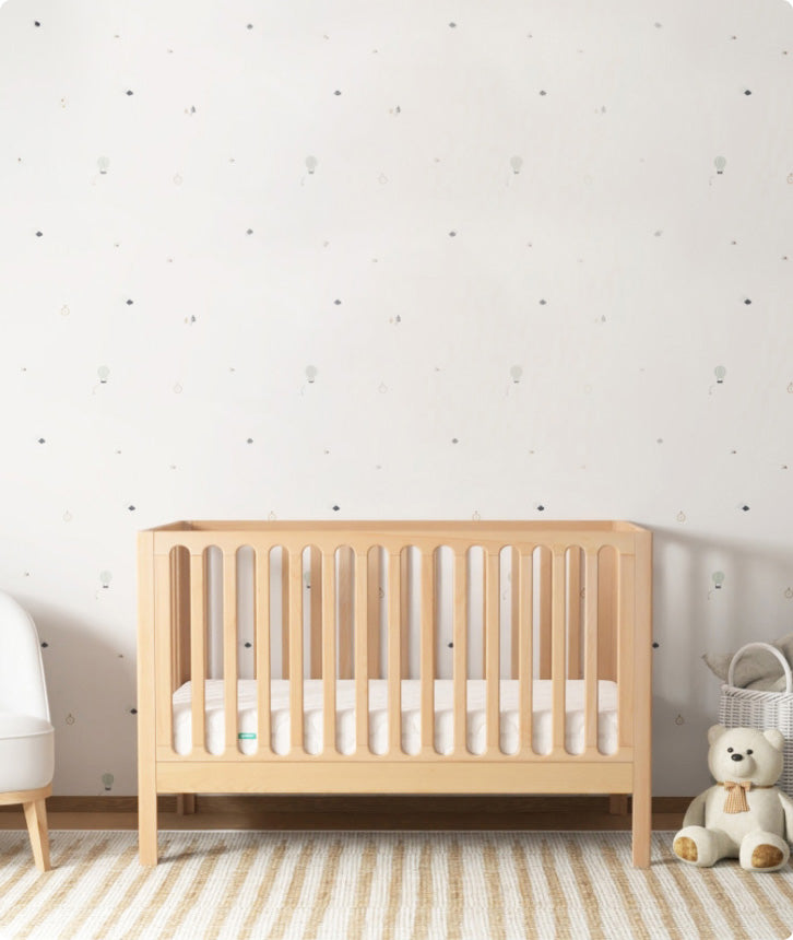 Light wood Newton crib centered in a nursery; teddy bear and basket fully visible on the right, with a partially visible white chair on the left.