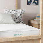 A white mattress is on a light-brown wooden bed frame.  The mattress has a light teal-colored wordmark that reads "newton" on it. A white pillow with a black polka dot pattern is positioned on the top of the bed above the mattress. Another white pillow is underneath the polka dot pillow.  A small wooden nightstand is visible in the background to the right of the bed.  A framed print of space imagery is on the wall behind the bed. The walls are a light beige or cream color.