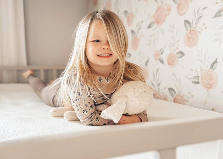 A young girl with blonde hair is lying on a white crib mattress.  She is wearing a light brownish-beige long-sleeved top with a subtle floral pattern and brownish-beige pants.  She is smiling and holding a light beige stuffed animal that resembles a dog or puppy.  The wallpaper behind her is a pale pastel pink and gray floral design. The crib is light gray-white.  The room appears to be a child's bedroom or nursery.