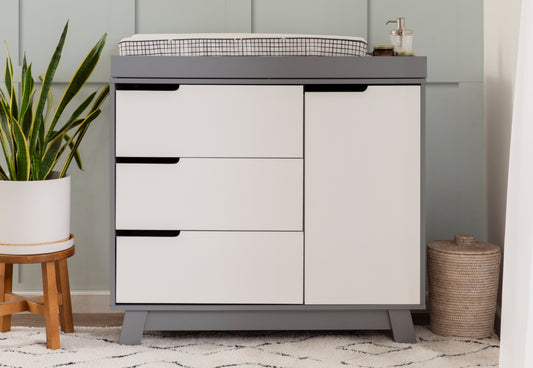 A modern changing table, set in a nursery featuring a plant and a woven basket, against a light-blue wall. 999-8601-GYW