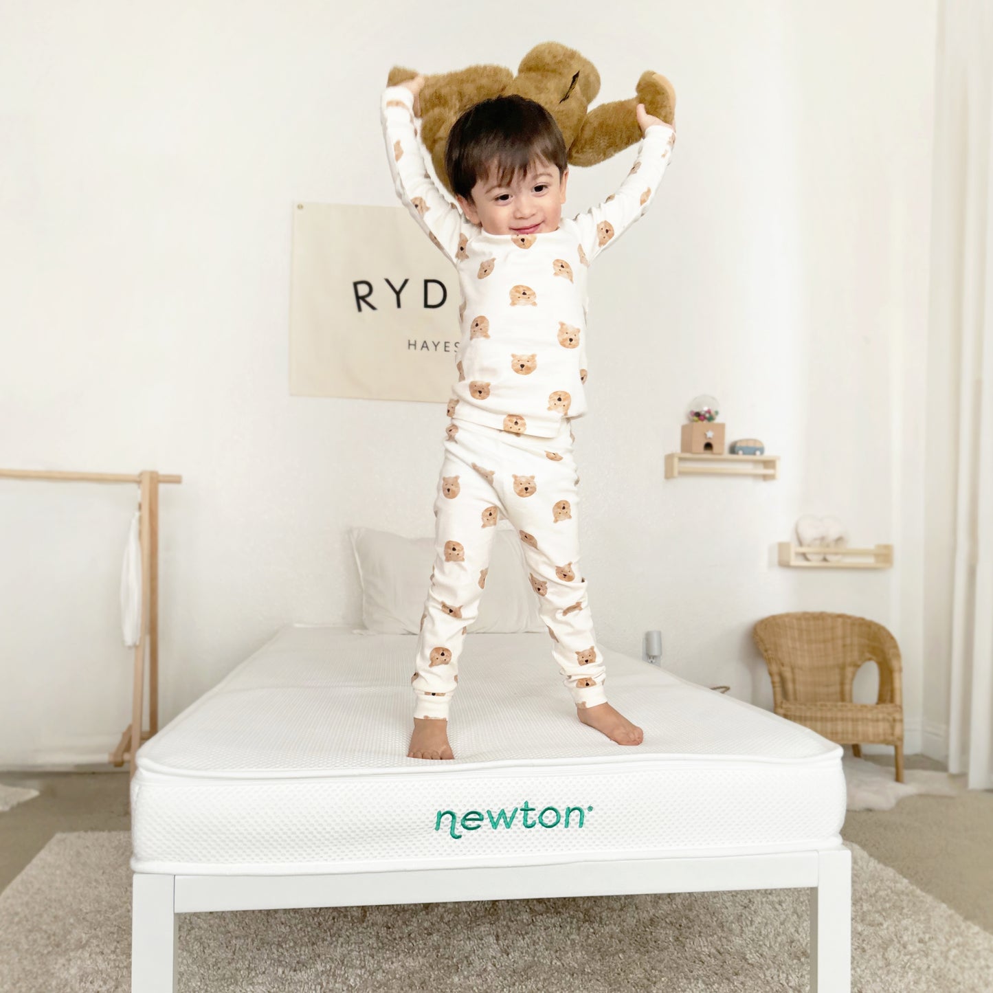 A smiling toddler in pajamas stands on a Newton Baby Kids' Twin Mattress, holding a plush teddy bear overhead. The softly lit room features minimalist decor and neutral tones.