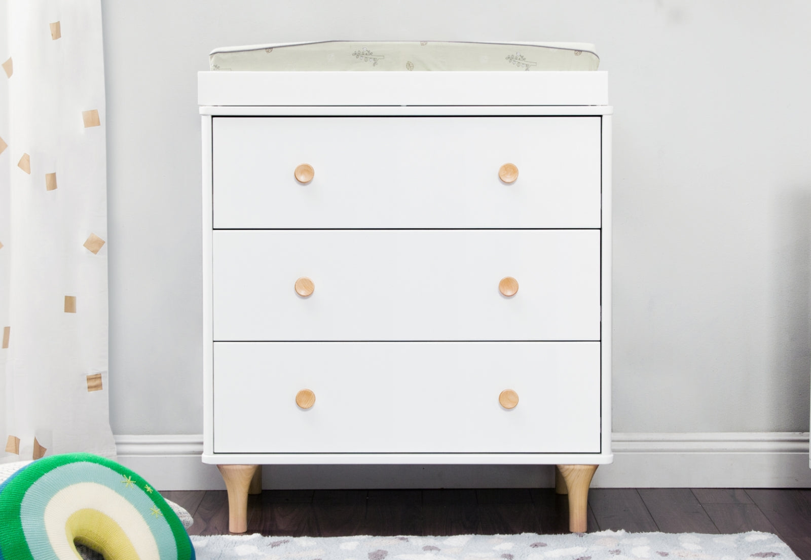 A white nursery dresser with three drawers and wooden knobs, positioned against a light grey wall, nearby a colorful toy.