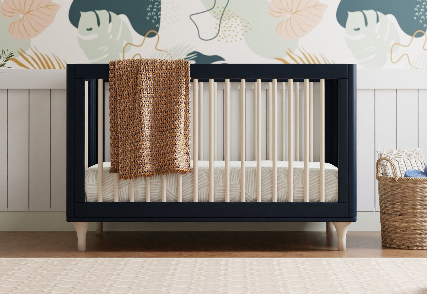 Dark blue baby crib with a knitted blanket draped over the side, positioned in a nursery room with a whimsical elephant-themed wallpaper and a wicker basket.