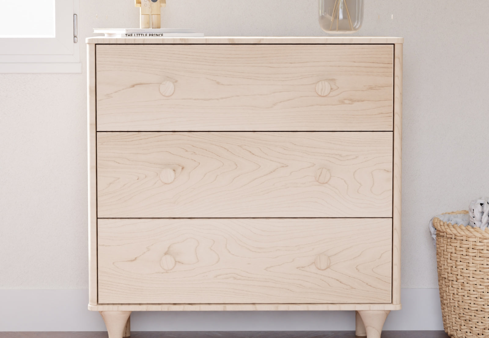 Modern light wooden nursery dresser with simple, angular handles in a clean, neutral-toned room, with a small wicker basket partially visible to the right.