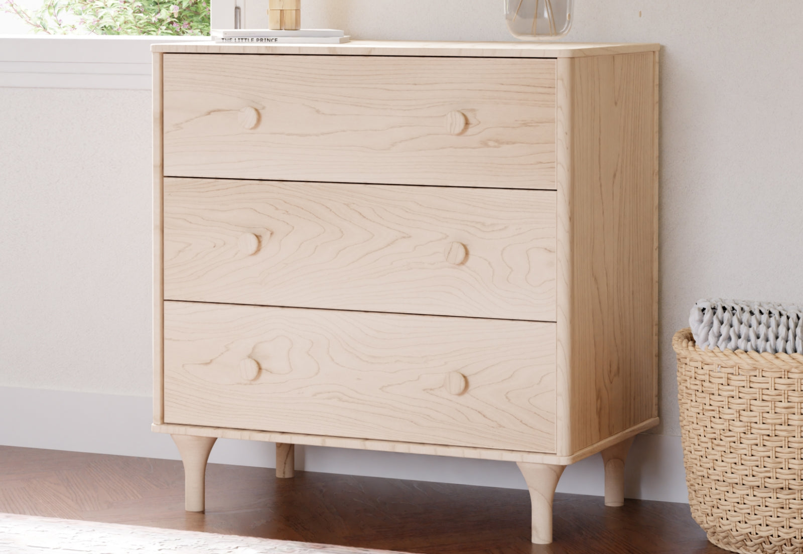 A minimalist wooden dresser featuring three drawers with circular wooden knobs, set against a light modern interior including a basket and a book titled "The Little Prince" on top.