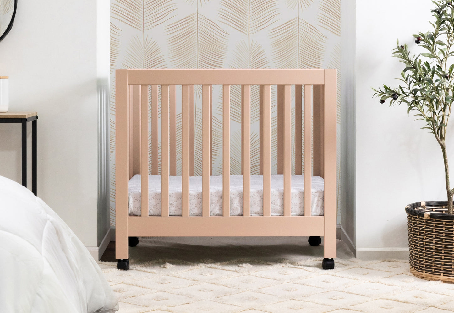 A modern light pink crib in a stylish bedroom with a neutral color palette, featuring a leaf-patterned wallpaper and a green potted plant. 999-8505-CAN