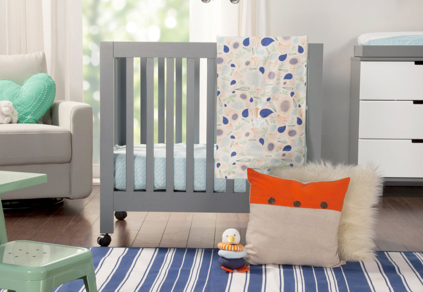 A cozy nursery featuring a modern gray crib, striped blue and white rug, colorful decorative pillows, and a playful toy on the floor. 999-8505-GRY