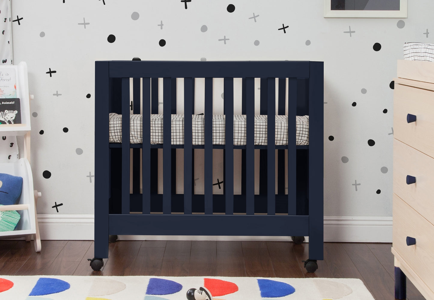 Navy blue baby crib in a nursery with playful black and white wall decals and a colorful circular patterned rug.