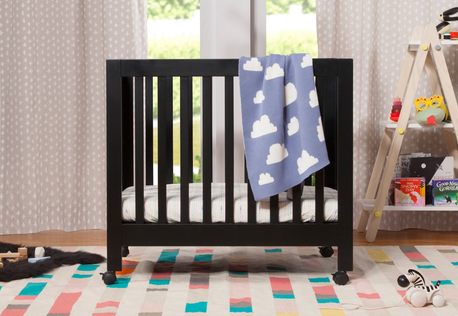 A cozy nursery setup featuring a dark wood crib with a blue cloud-patterned blanket, colorful striped rug, and a toy shelf with children's books and toys. 999-8505-BLK
