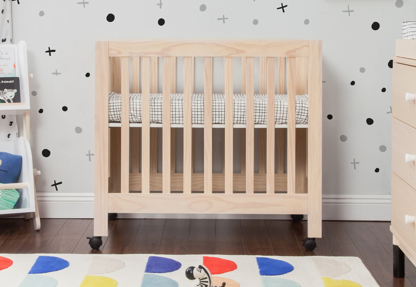 A sturdy wooden crib in a nursery with geometrically patterned bedding, set against a playful wallpaper with black and grey crosses and dots, next to a colorful, abstract rug. 999-8505-WNA