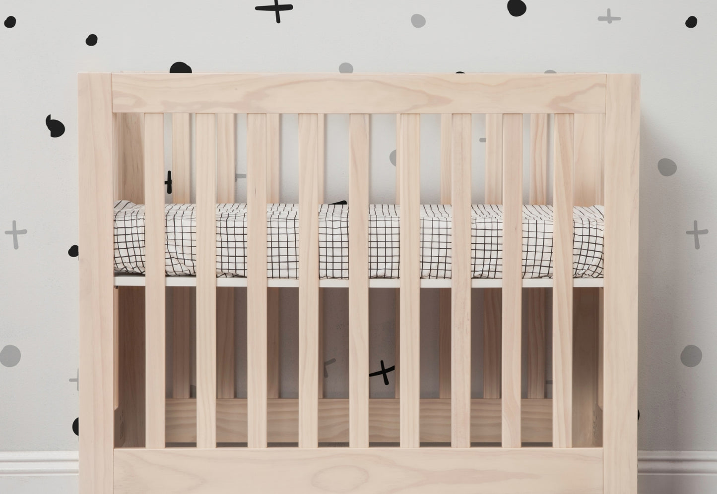 A modern wooden crib in a nursery with a patterned mattress and stylish black and gray dot and cross wall decor.