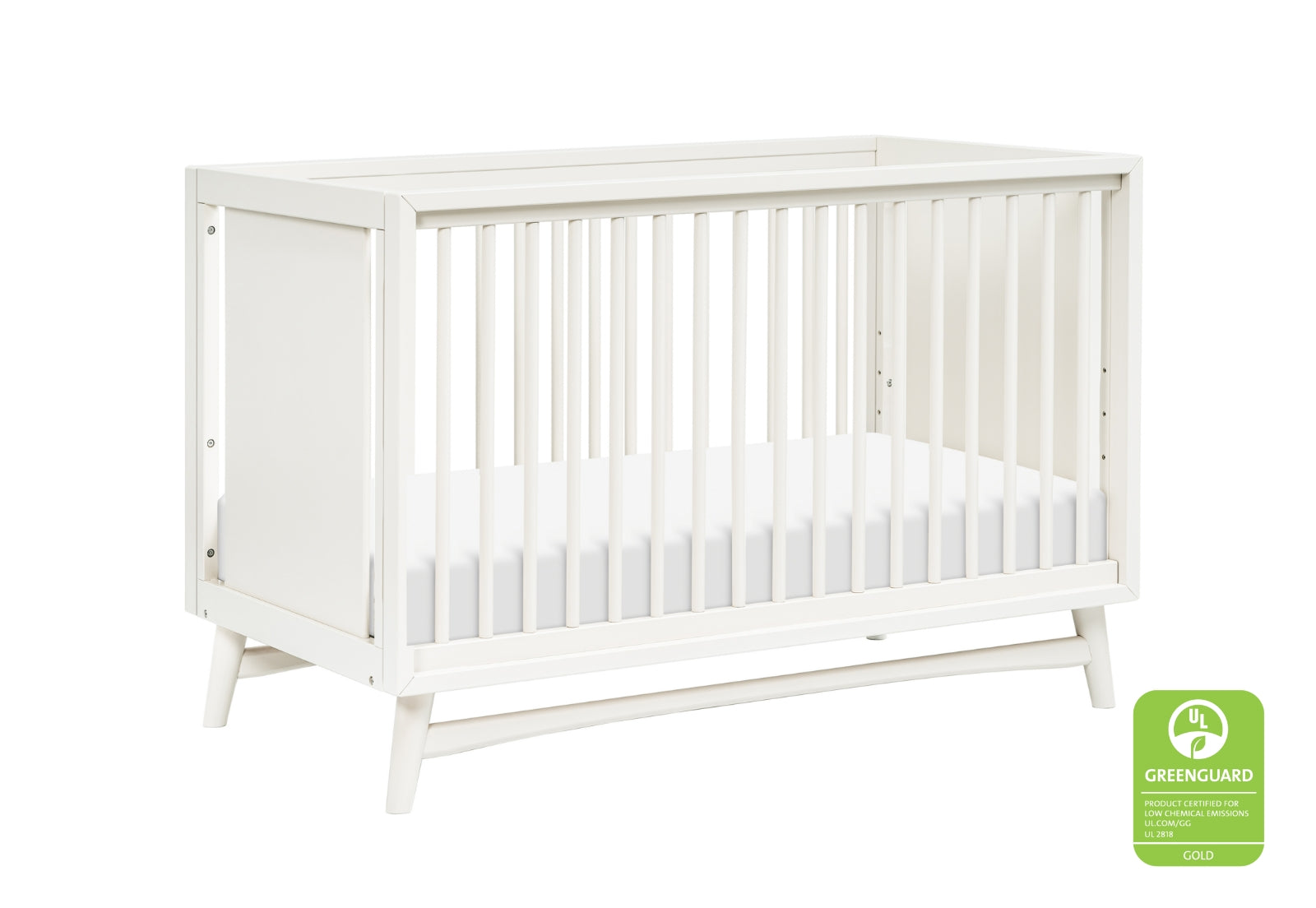 The Babyletto Peggy Convertible Crib, featuring white wood, slatted sides, and angled legs in a mid-century style, is shown with a plain white mattress and a Greenguard Gold certification badge in the corner.