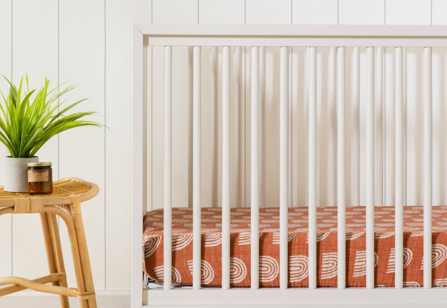 The Babyletto Peggy Convertible Crib with a rust sheet featuring white rainbows stands beside a wicker table holding a green potted plant and two jars, creating a cozy modern nursery against a white paneled wall.
