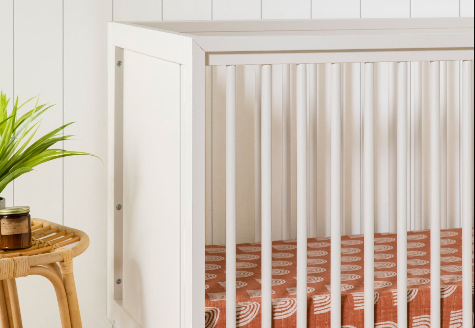 A modern nursery features the Babyletto Peggy Convertible Crib in white with vertical slats and a rust crib sheet with white rainbows. A woven side table stands nearby, topped with a green potted plant and a small jar.