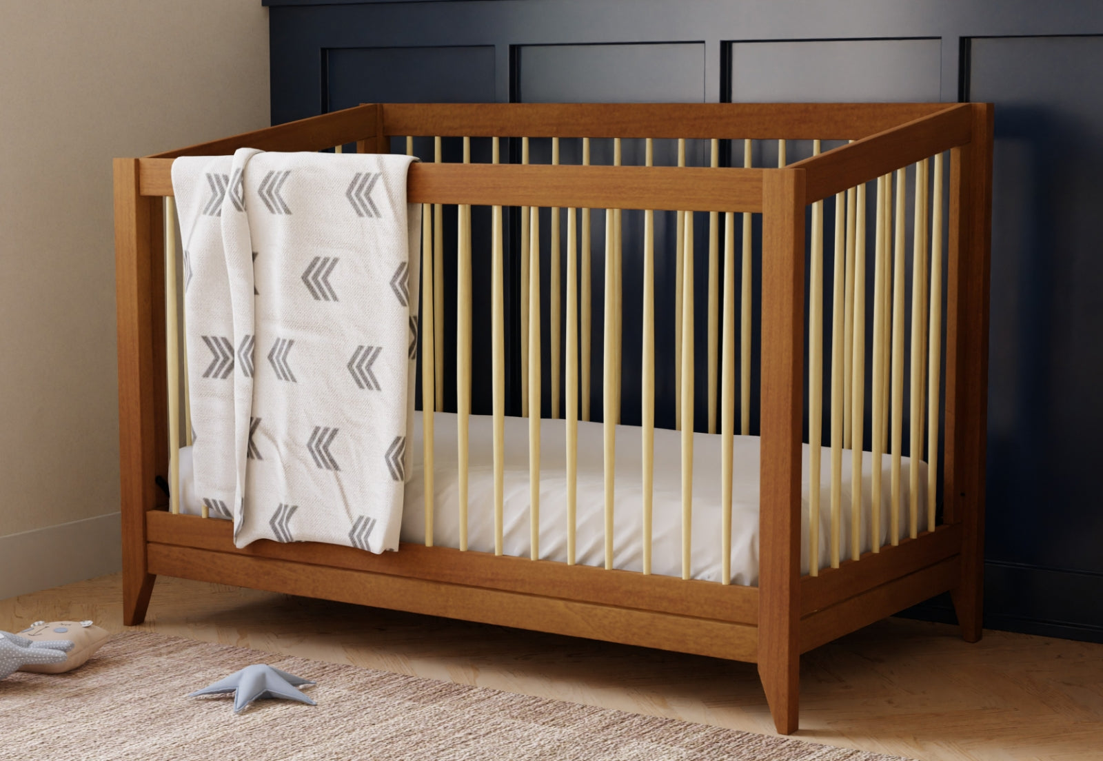 The Babyletto Sprout Convertible Crib, featuring light-colored vertical slats, stands on a light rug with a white blanket adorned with gray arrows, set against a dark blue paneled wall—modern nursery furniture at its best.