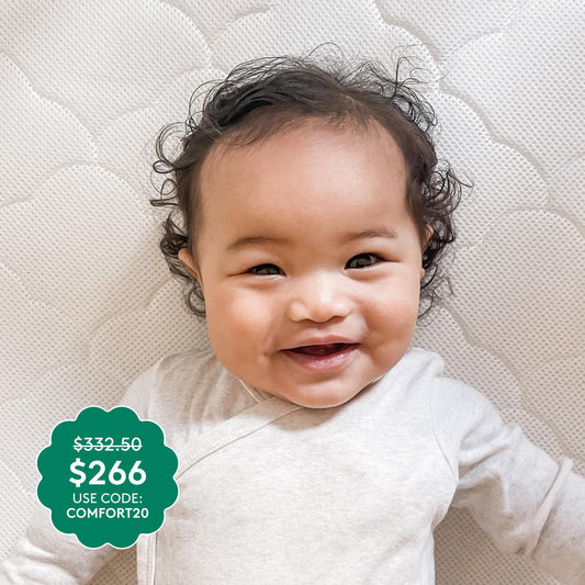 A smiling baby in a light onesie lies on a textured white surface atop the ultra-breathable Newton Baby Waterproof Crib Mattress. A green label shows $266 (was $332.50) with promo code COMFORT20.