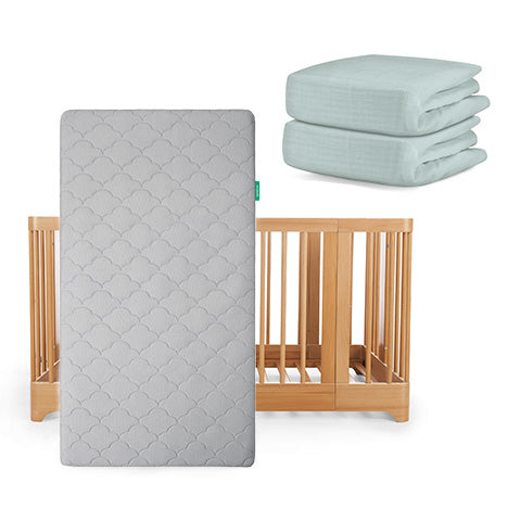 The Newton Baby Nest Convertible Crib has vertical slats and is paired with an Original Mattress in a gray pattern and two folded light blue Breathable Organic Cotton Sheets.