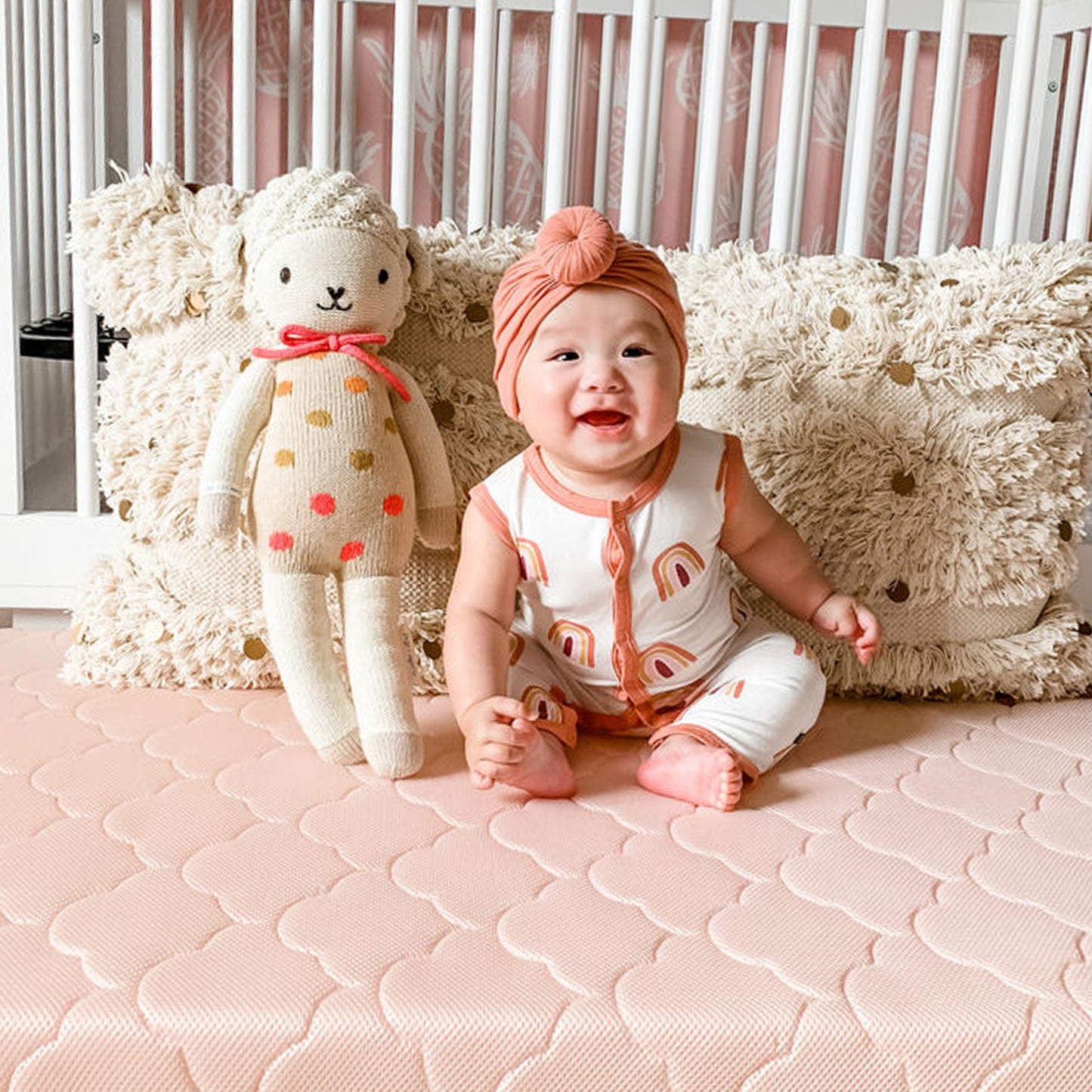 cheerful baby sitting on newton pink mattress with stuffed animal and pillows