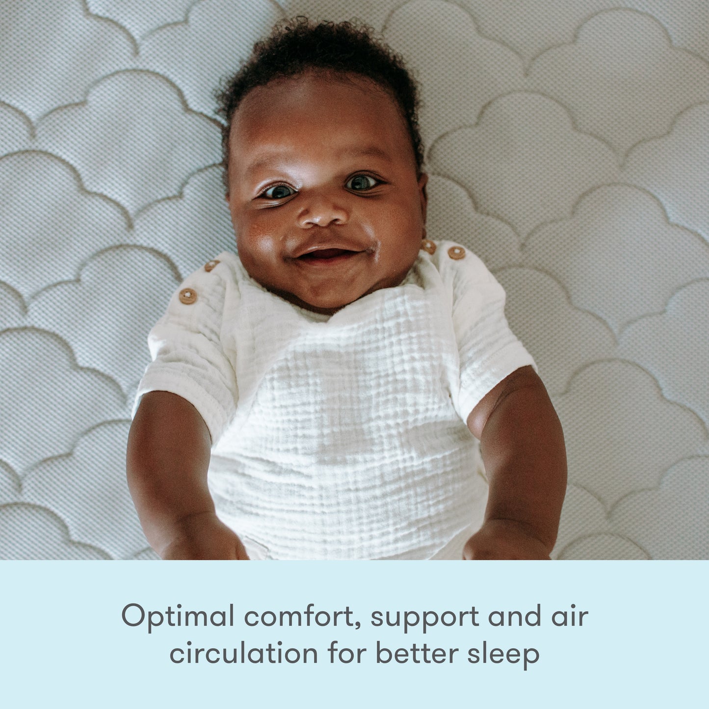 A cheerful baby laying on a gray, cloud-patterned mattress, smiling at the camera. with text "optimal comfort support and air circulation for better sleep"