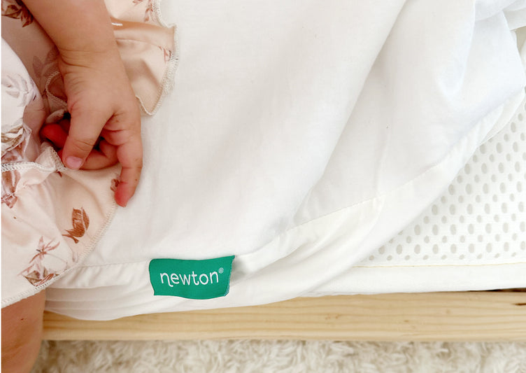 ALT text: Close-up of a baby's hand gently resting on a Newton mattress pad with a visible Newton tag.