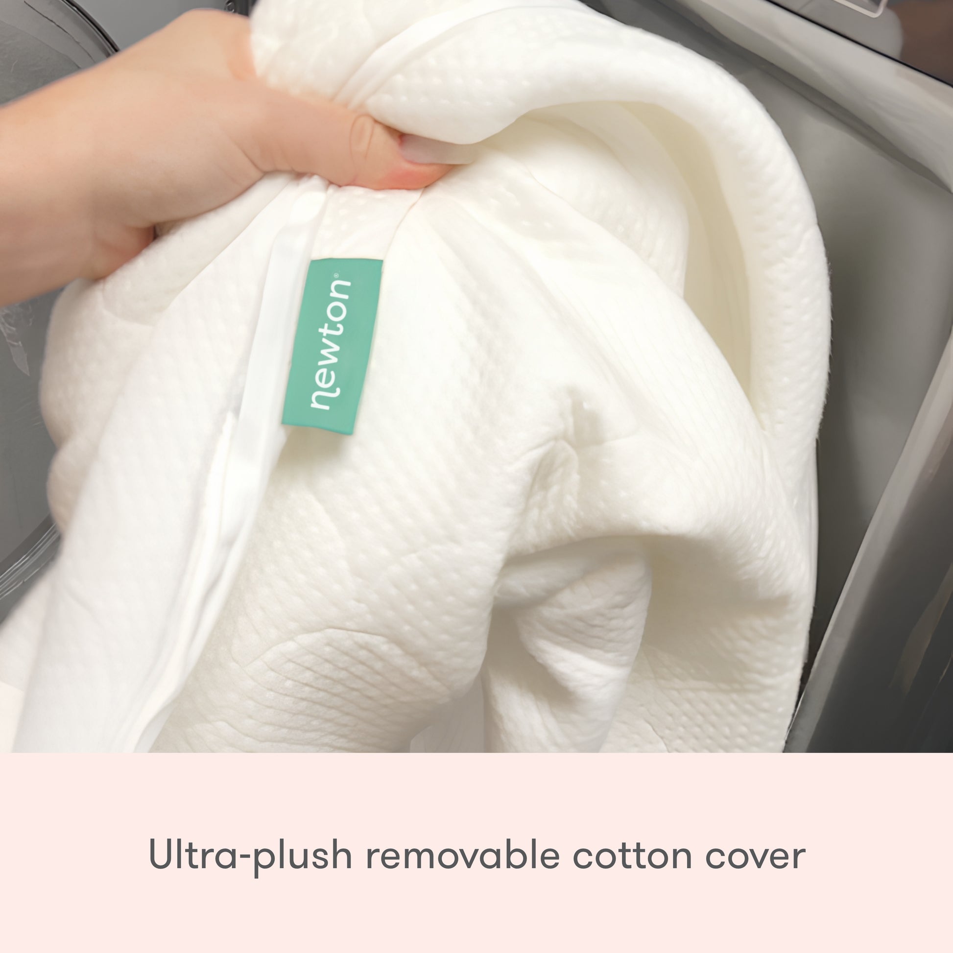 A hand holds The Pregnancy Pillow’s ultra-plush, white cotton cover with a green Newton Baby tag beside a washing machine. Text: Removable, soft cotton cover for The Pregnancy Pillow by Newton Baby.