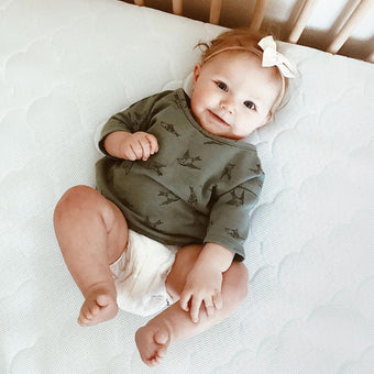 A baby girl in a green shirt lies down happily on a Newton mattress.