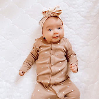 A baby girl in a brown polka dot onesie, smiling and lying on a Newton mattress.