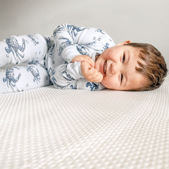 A baby lying on a white newton mattress, playfully resting his hands on his face, looking adorable and content.