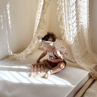 A baby girl sitting on a newton mattress with a delicate white canopy, smiling and surrounded by soft pillows.