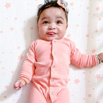 a baby wearing a peach-colored long-sleeved onesie. The onesie is a simple design with small, light-colored buttons down the front. The baby is lying on a crib sheet with a pale peach star pattern. The baby appears happy and is wearing a peach-colored bow in its hair.